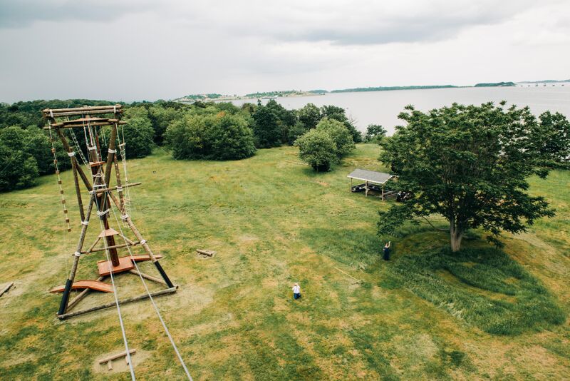 The image shows an aerial view of an outdoor adventure course. A tall wooden structure dominates the foreground, likely a climbing or zip-lining platform. The course is set in a grassy field with trees and a body of water in the background. The sky is overcast.
